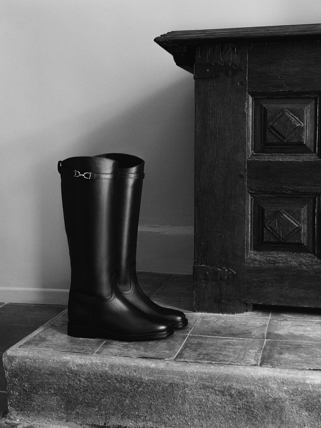 Black leather riding boots next to a wooden cabinet, in black and white — Massimo Dutti Fall Treasures