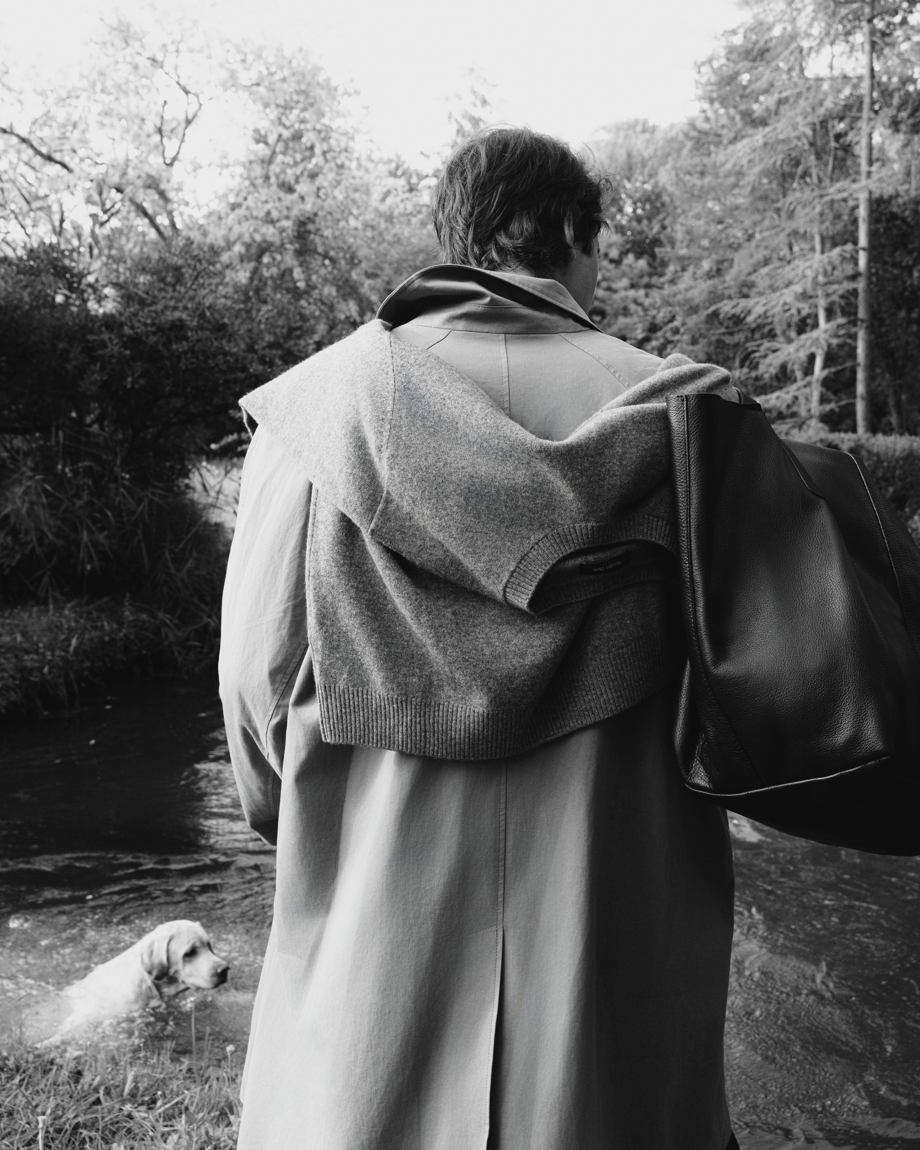 Man from behind in a trench coat, sweater over his shoulders and tote bag by a river, in black and white — Massimo Dutti Fall Treasures