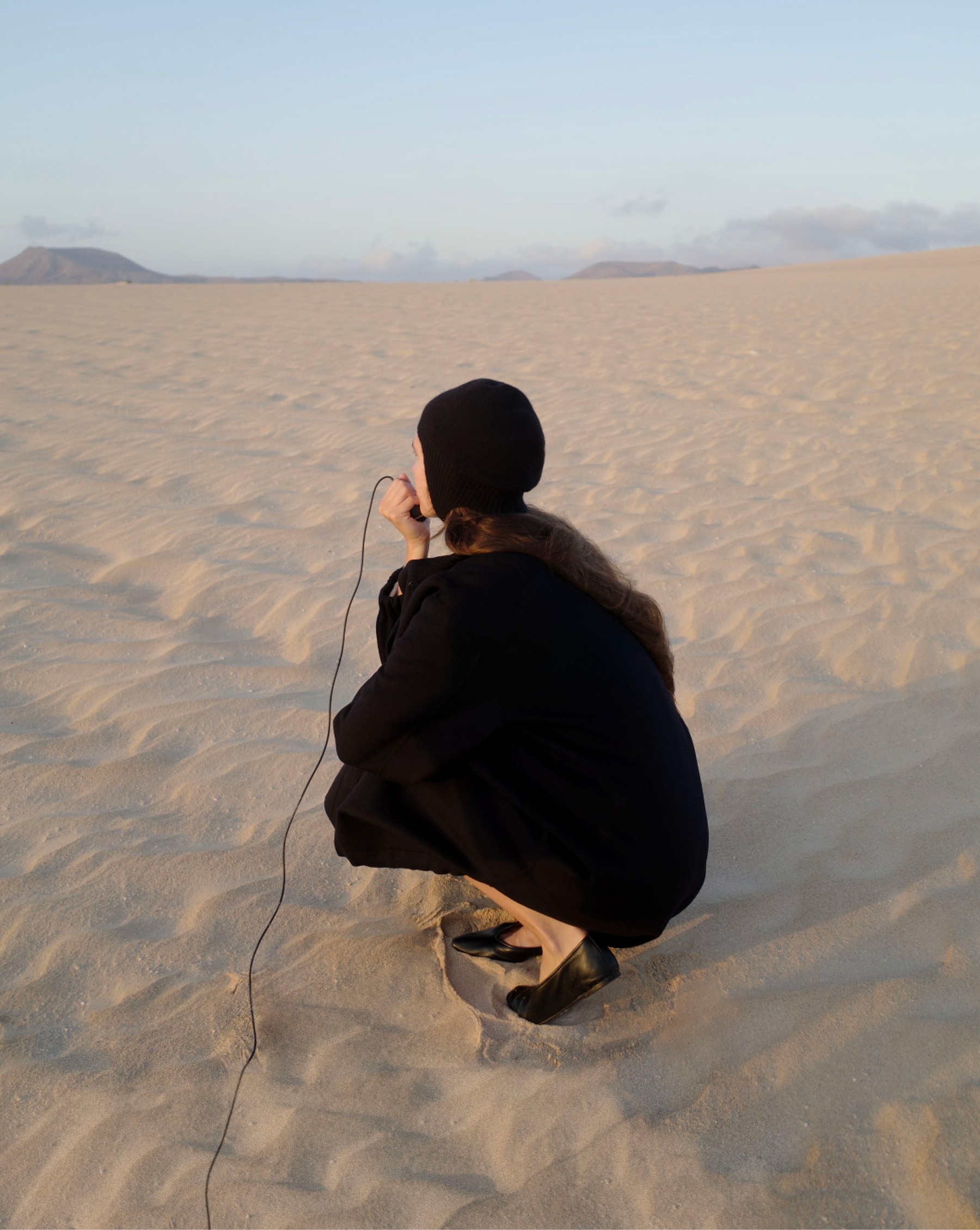 Model crouching in a black coat on the dunes, Fuerteventura — Massimo Dutti