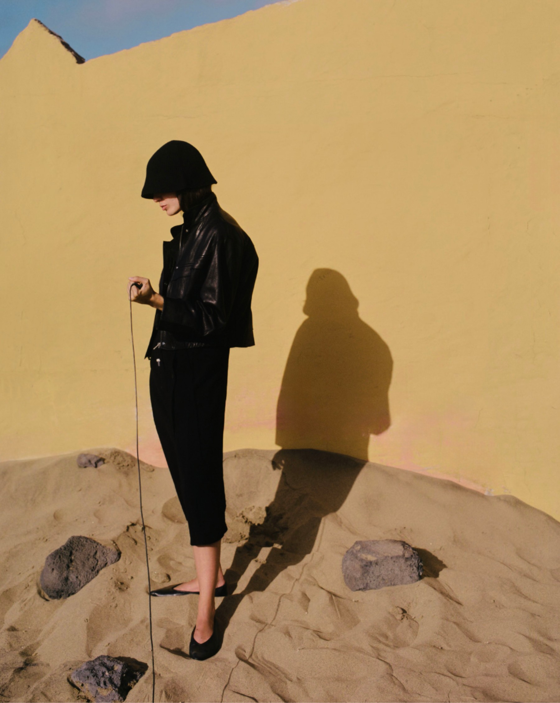 Model in a bucket hat and short black leather jacket on the sand, before an ochre wall — Massimo Dutti