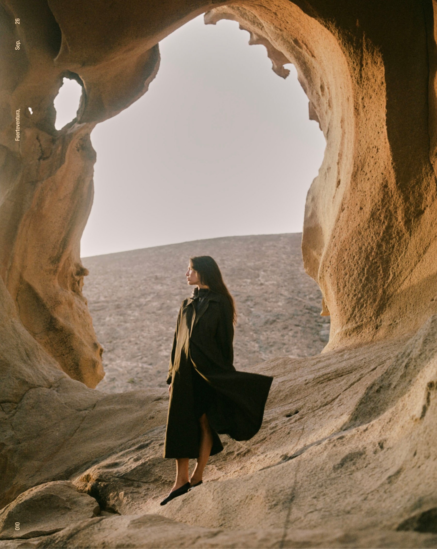 Model in a dark trench coat beneath an arch of eroded rock, Fuerteventura — Massimo Dutti