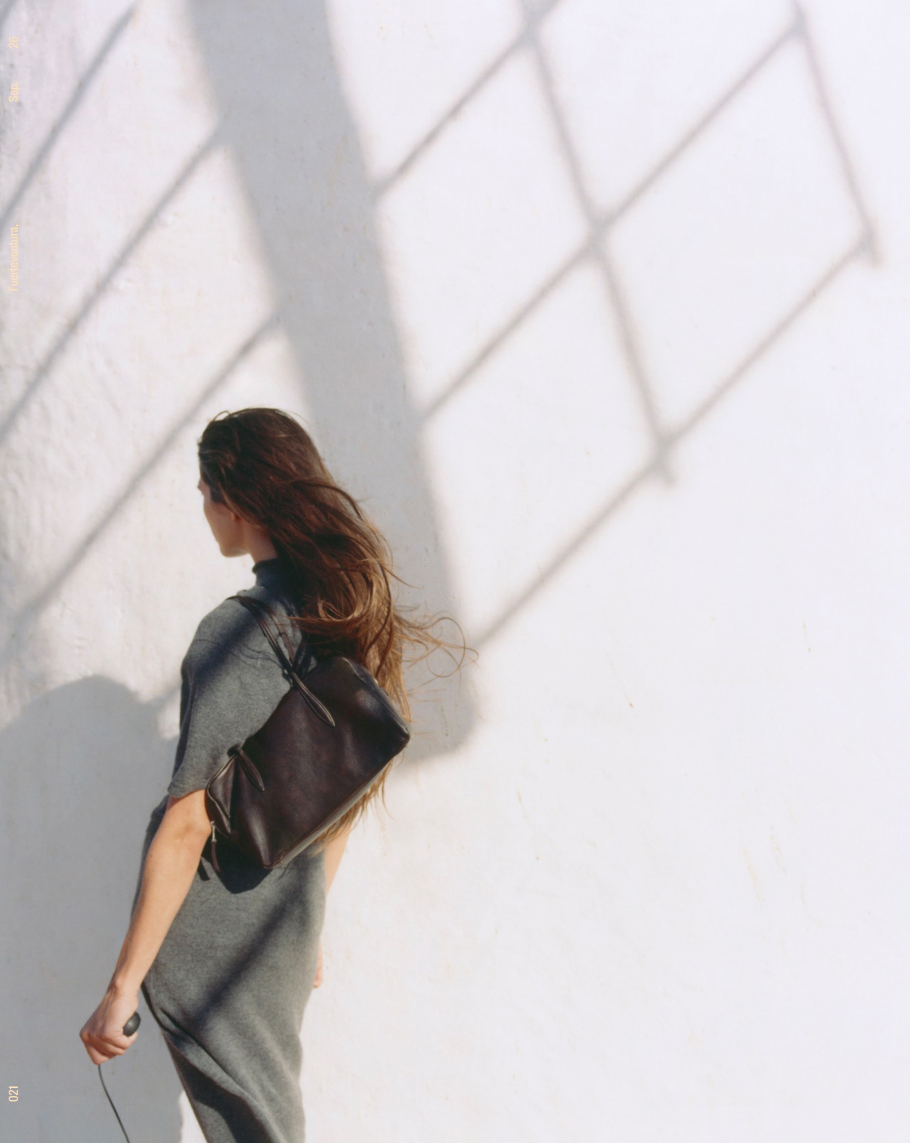 Model in a grey knit dress with a black mini satchel bag before a white wall with shadows — Massimo Dutti