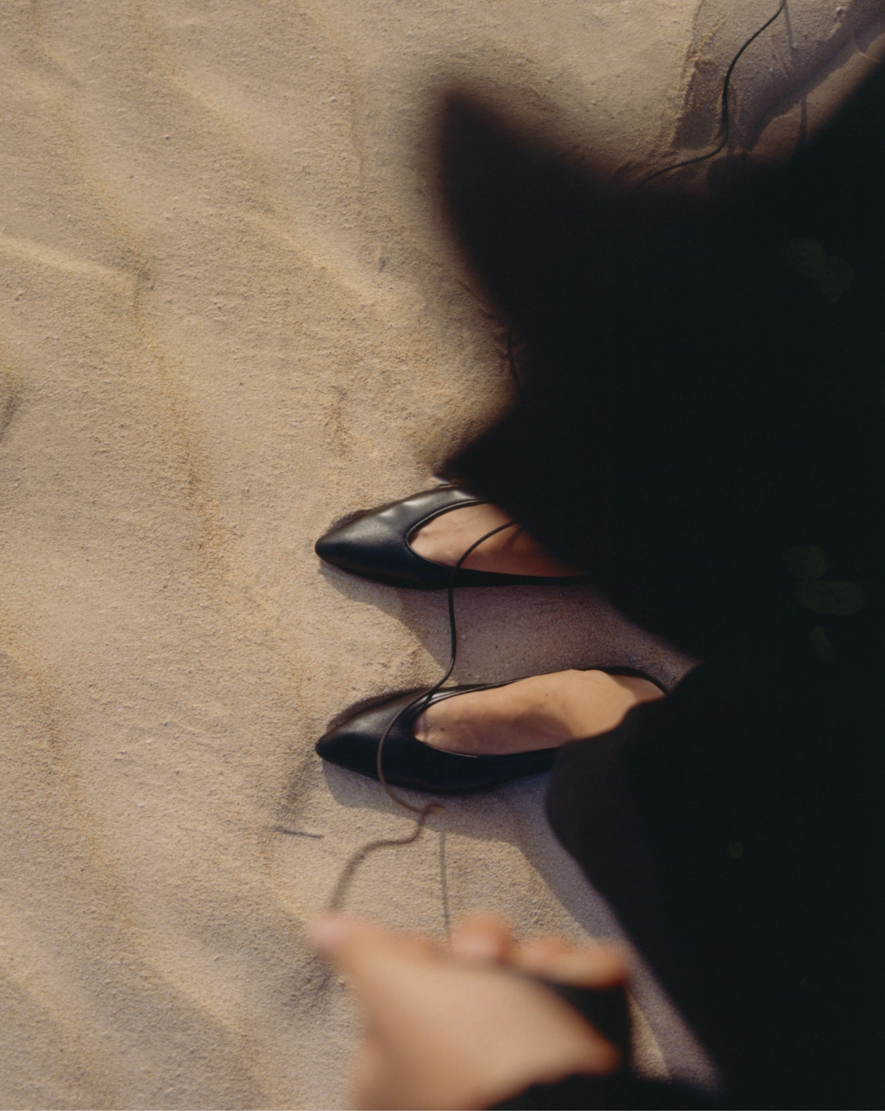 Black leather ballet flats on the sand, with the model’s shadow — Massimo Dutti